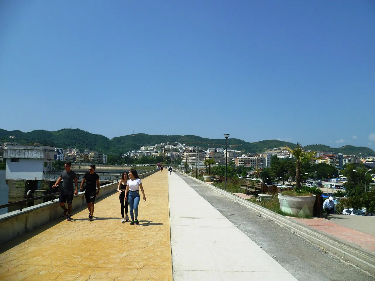 People walking the lakeside promenade of the Grand Park in Tirana, with the city and green hills behind