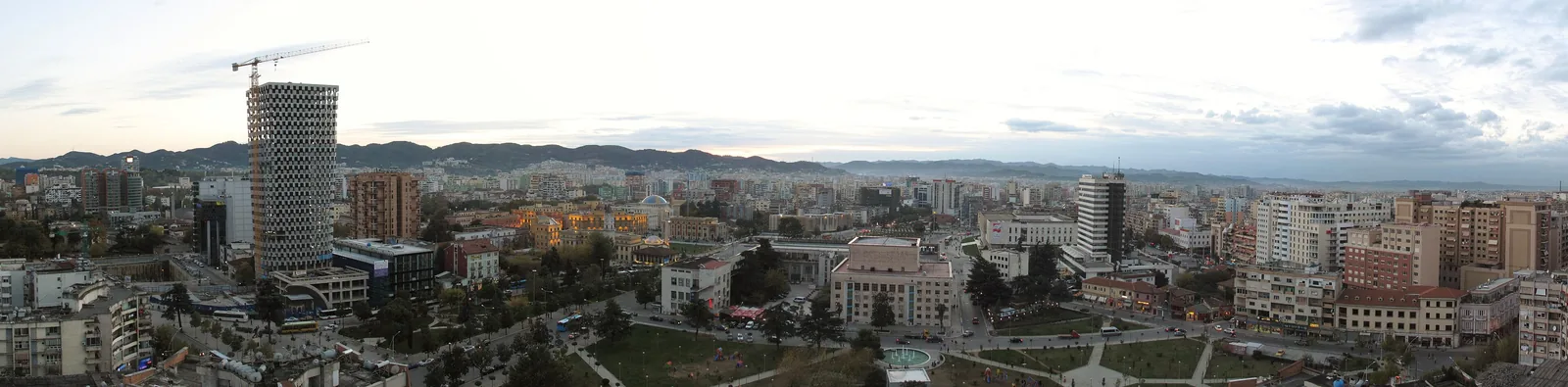 A wide panorama across central Tirana at dusk, the Pyramid-area towers and city rooftops spread out below the surrounding hills