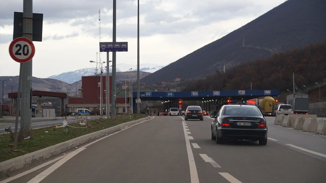 Cars in two lanes rolling toward the blue canopy of the Vërmicë border post on the Kosovo side of the Morinë crossing, a mountain rising behind it