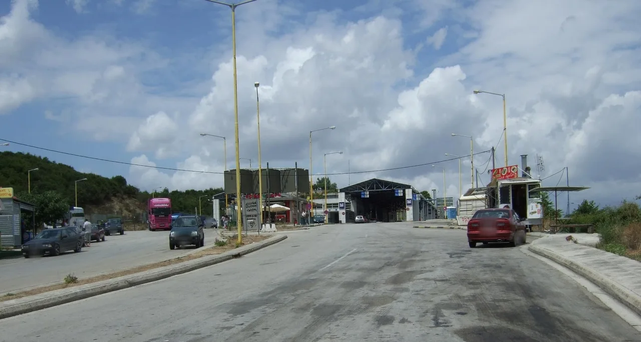 The Kakavia border crossing under a cloudy sky, with lamp posts, roadside kiosks, a lorry and cars heading toward the inspection canopy