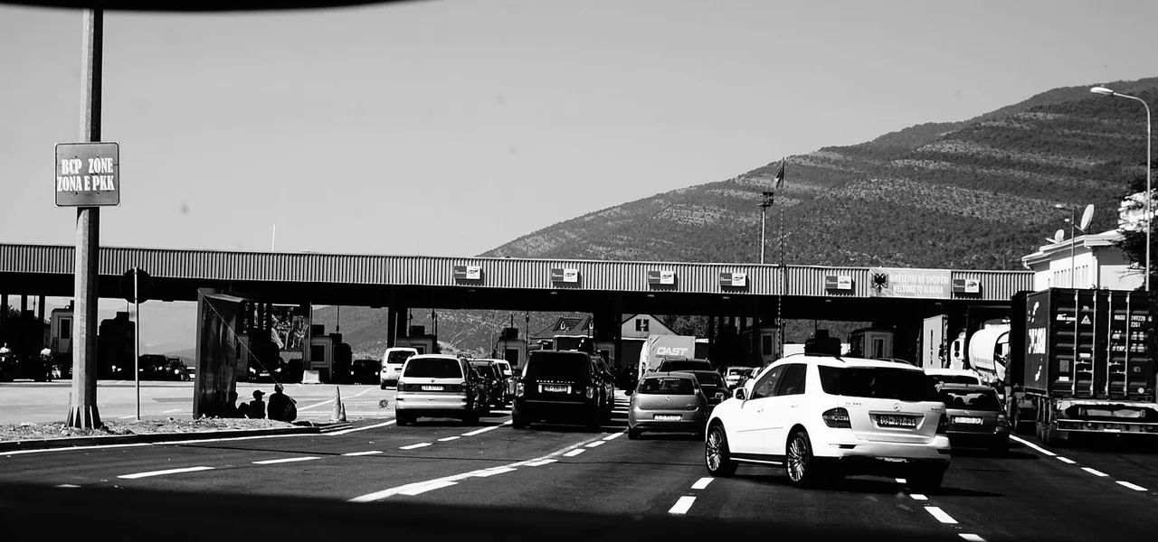 Black and white view through a windscreen of cars and lorries queued in several lanes under the Welcome to Albania canopy at Morinë