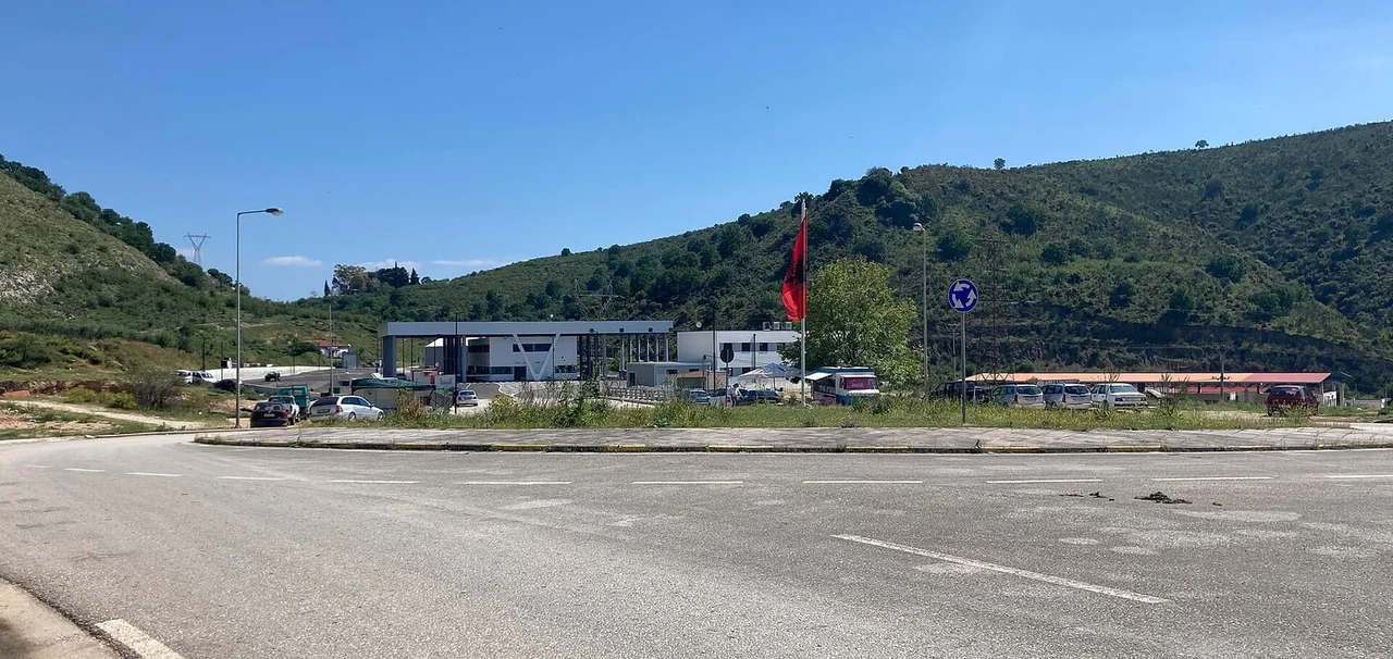 The low white buildings and canopy of the Qafë Botë border post on the Albanian side, an Albanian flag by the road and green hills behind