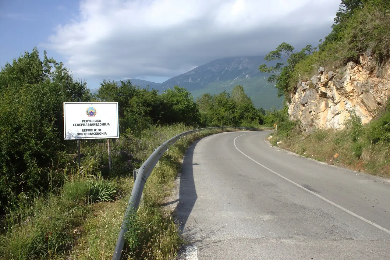 A curving road past a Republic of North Macedonia sign in Macedonian and English, with a rock cutting on the right and a mountain in cloud ahead