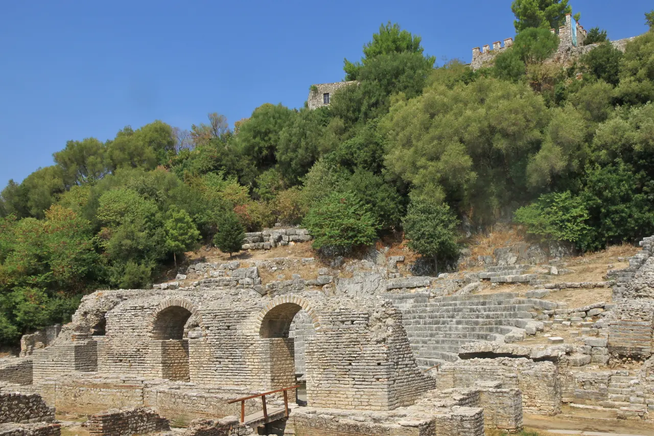 The ruins of the ancient theatre at Butrint among trees, with the old fortress wall behind