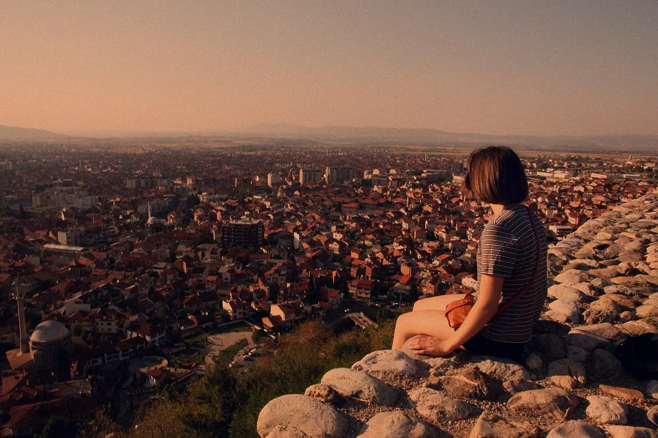 A visitor sitting on the walls of Prizren fortress at sunset, looking out over the red roofs of the old town
