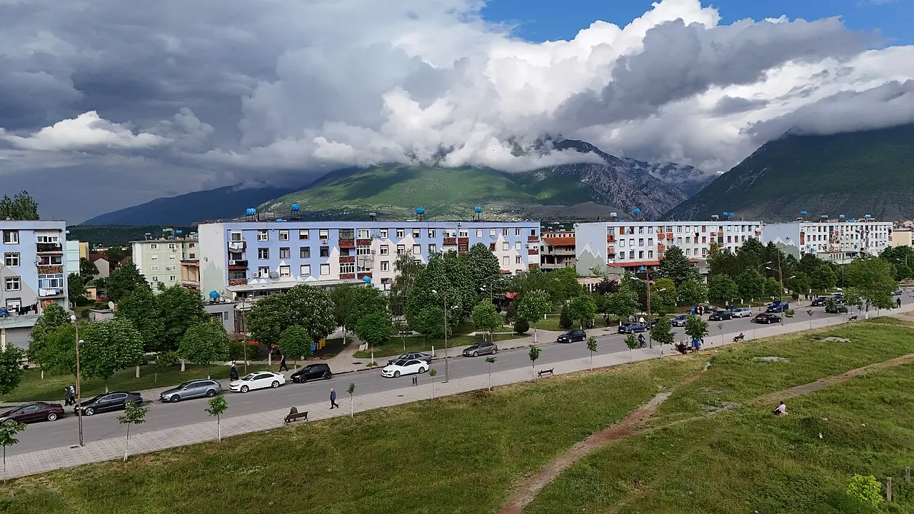 Aerial view of Kukes town's apartment blocks and a park-lined boulevard below the Alps, under a cloudy sky