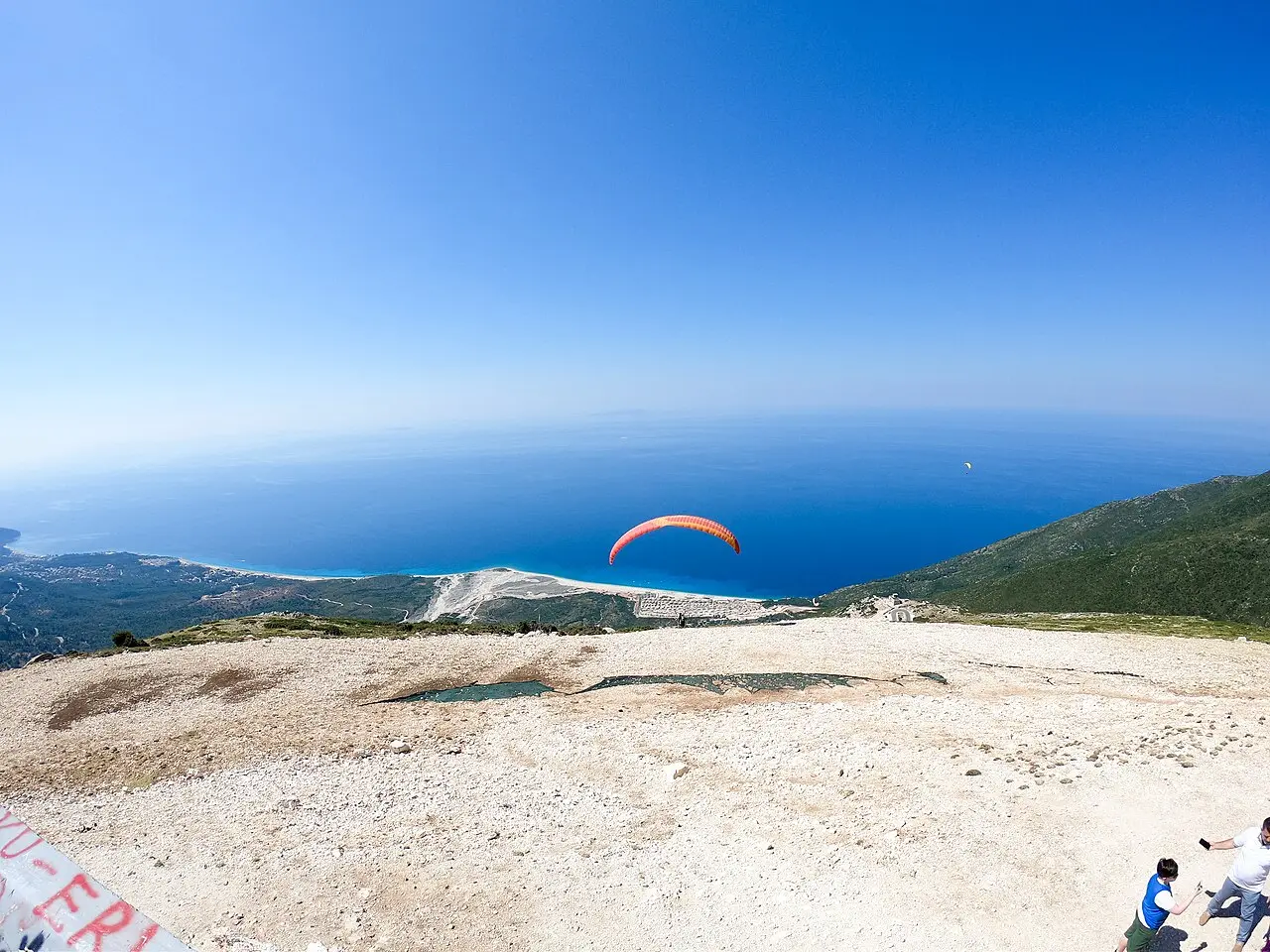 A paraglider launching from the Llogara heights over the turquoise Riviera coastline and the Ionian Sea