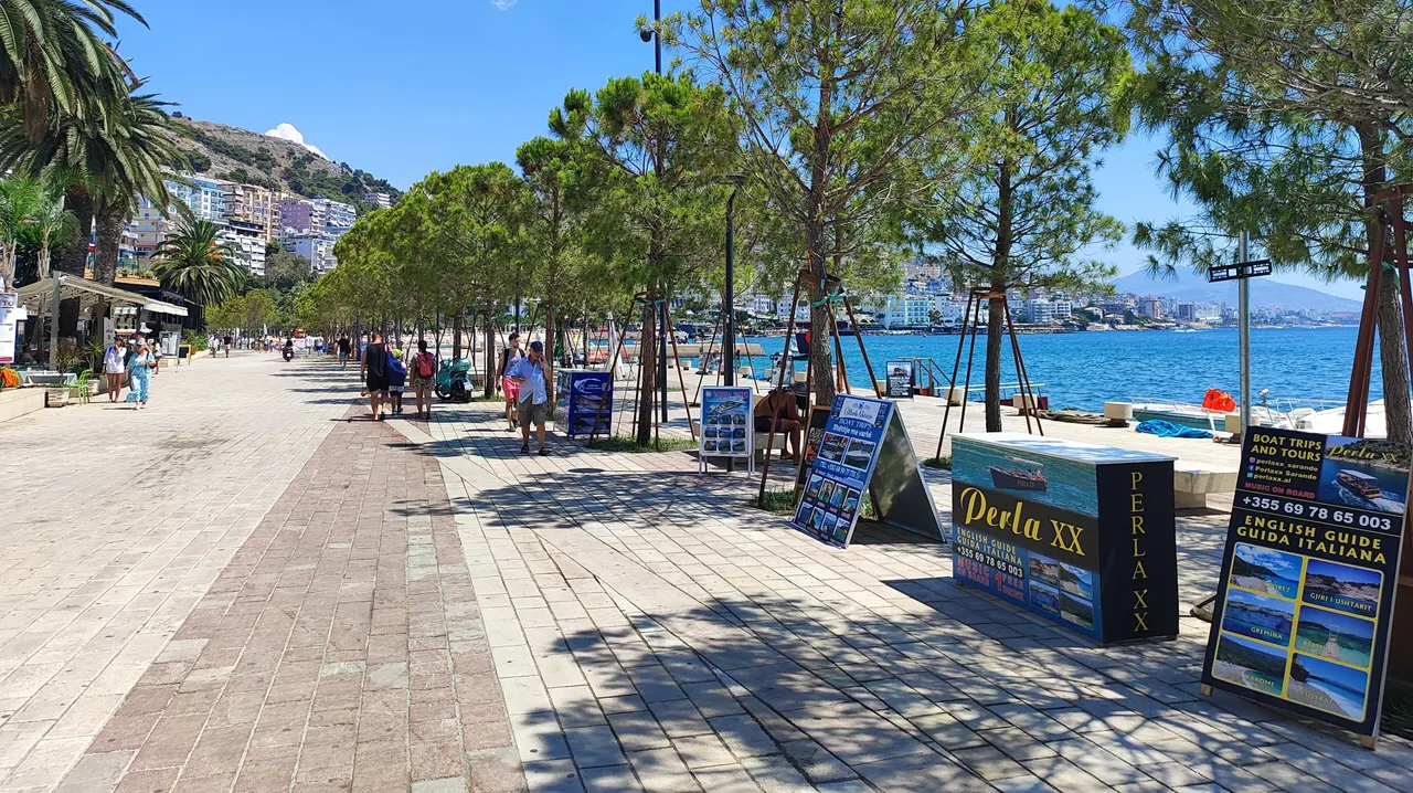 People walking the tree-lined seafront promenade in Saranda on a bright summer day, with the bay and town behind