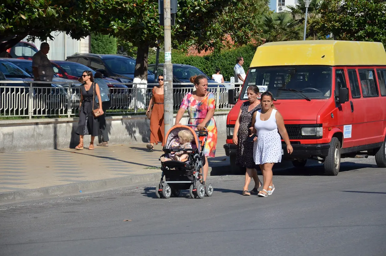 Women crossing a sunny street in Shkoder beside a red furgon van with a destination card in its windscreen
