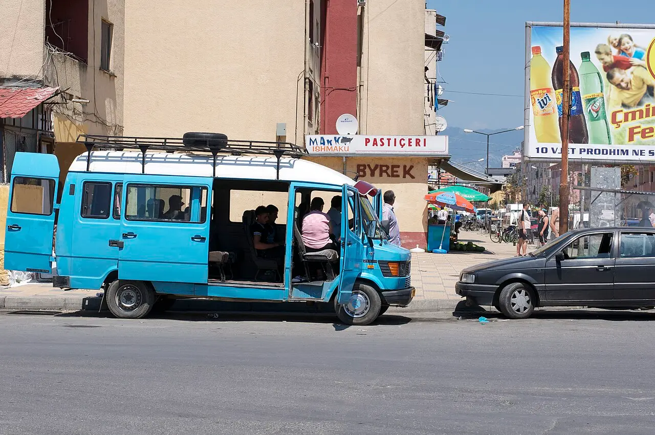 A blue Albanian furgon minibus with passengers parked on a town street beside a bakery