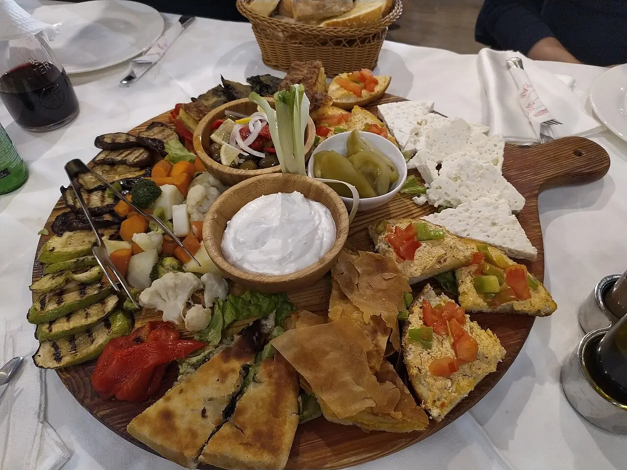 A large wooden board of grilled vegetables, feta, byrek pastry, dips and bread with a glass of red wine in an Albanian restaurant
