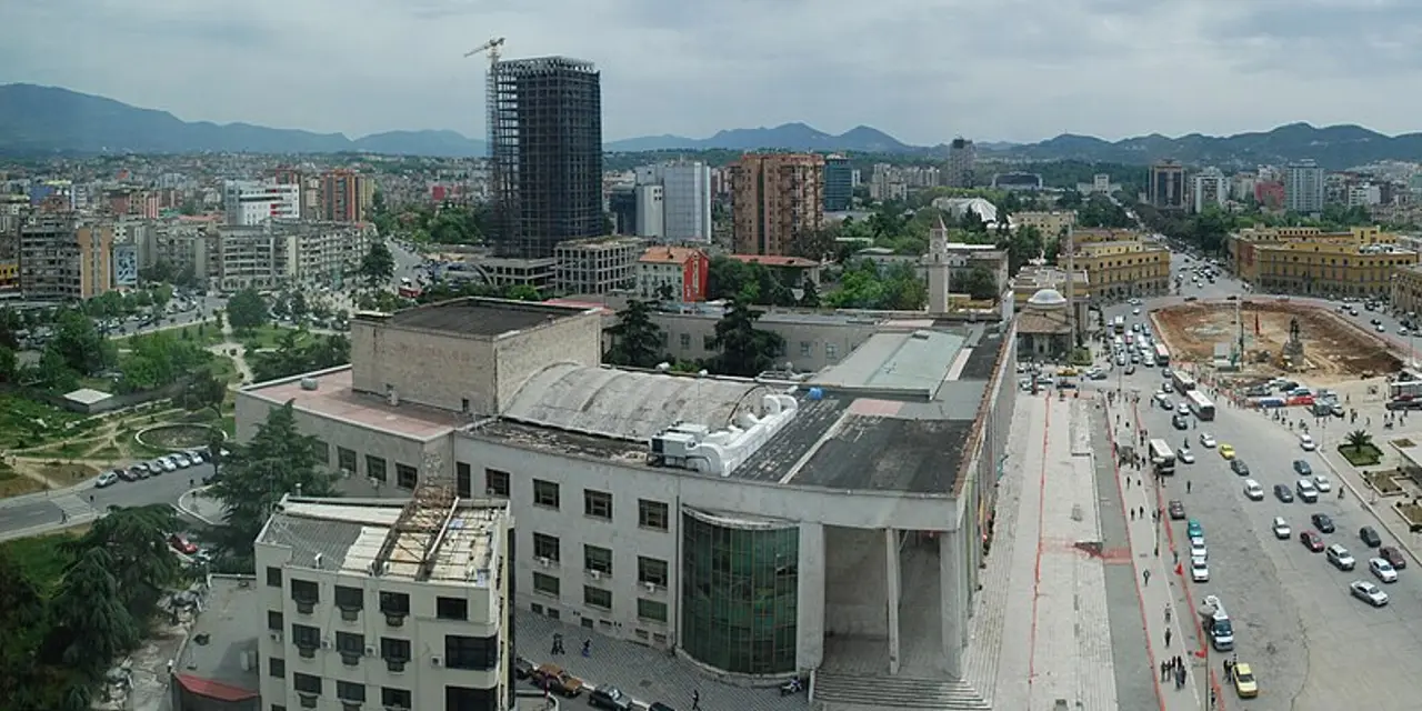 A rooftop view over central Tirana with mid-rise blocks, the clock tower and green hills ringing the city