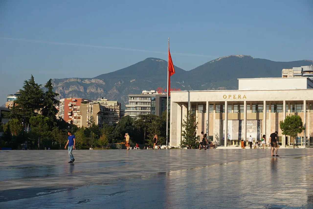 The Opera facade of the Palace of Culture on a wet paved square with the Tirana hills behind it