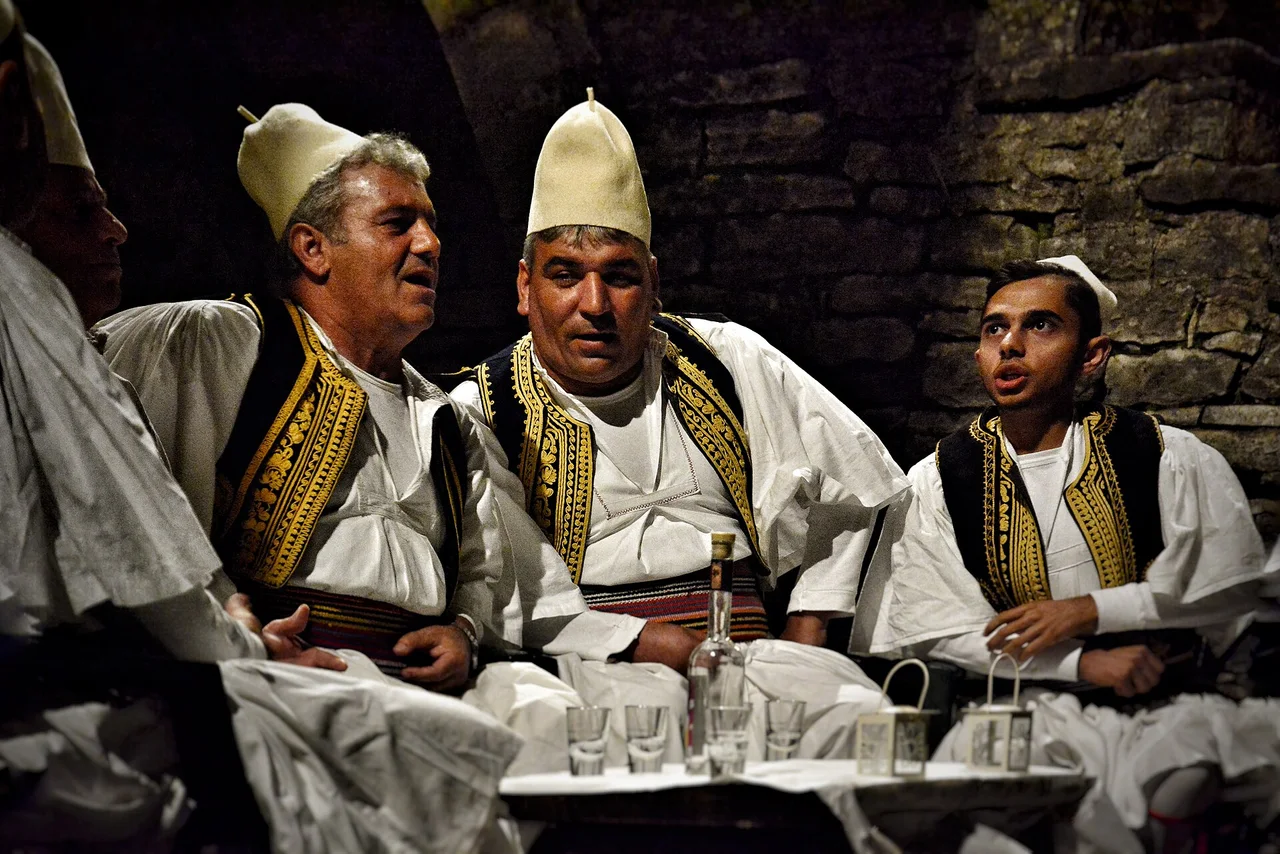 Three men in white felt caps and gold-braided waistcoats singing around a table with a bottle of rakia