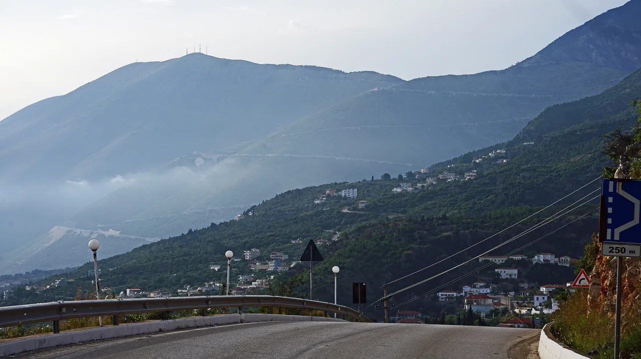 The coast road south of Vlora winding down through the Riviera hillside villages toward the Ionian, with the Ceraunian Mountains behind