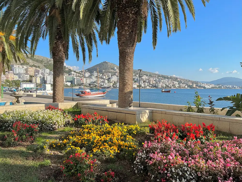 The Saranda seafront framed by palms and flower beds, with the bay, a moored boat and the town climbing the hill behind
