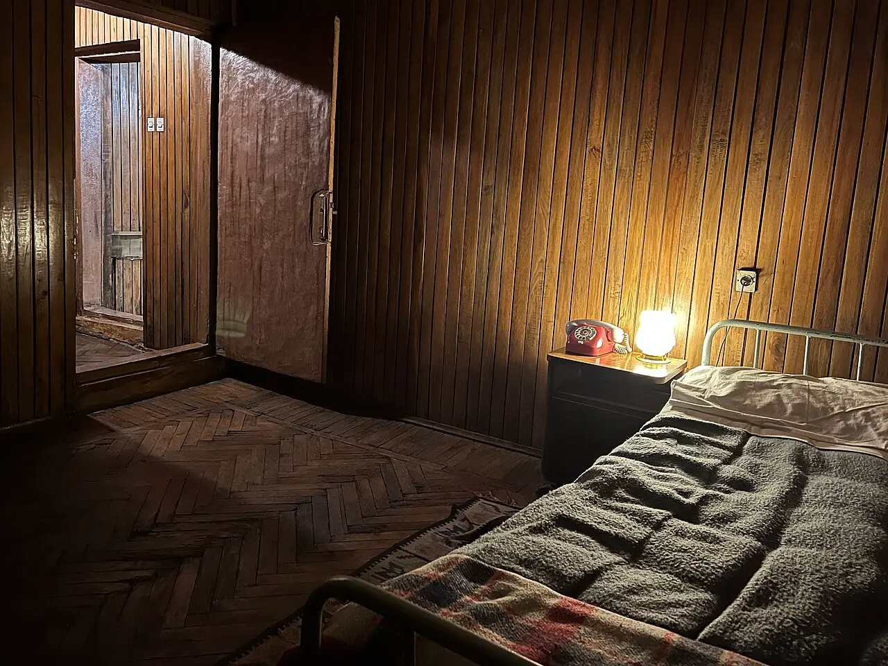 A wood-panelled bunker room with an iron bed and a red telephone lit by a bedside lamp