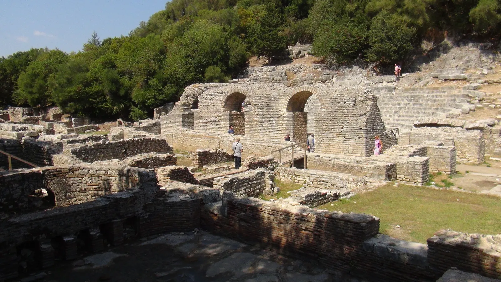 Stone ruins of the ancient city of Butrint with arched Roman walls and the theatre seating set against a wooded hillside