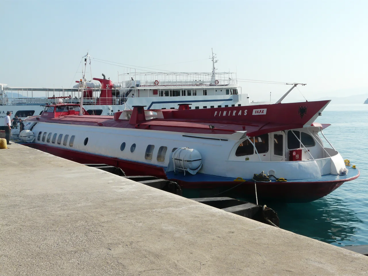 A red and white Finikas passenger boat moored alongside a concrete quay, with a larger ferry behind it