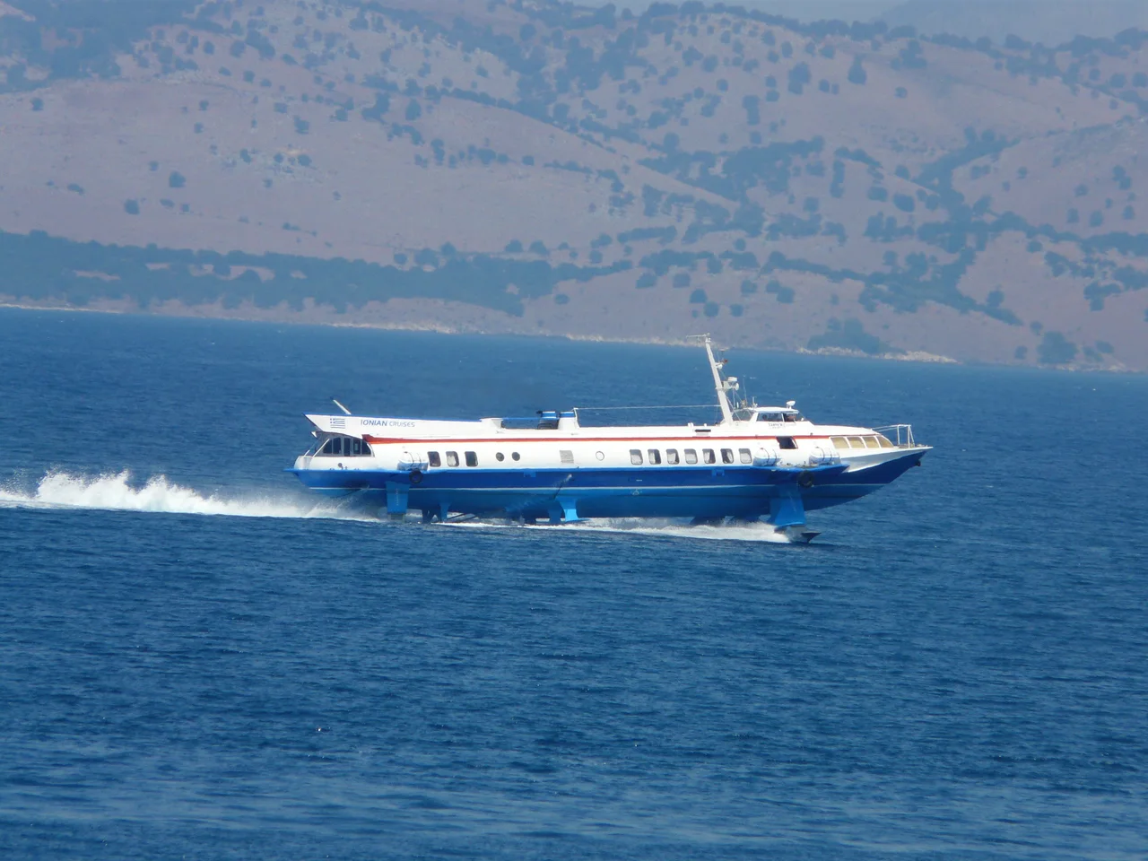 A white and blue hydrofoil speeding across the deep blue channel between Corfu and Albania, with hazy mountains behind