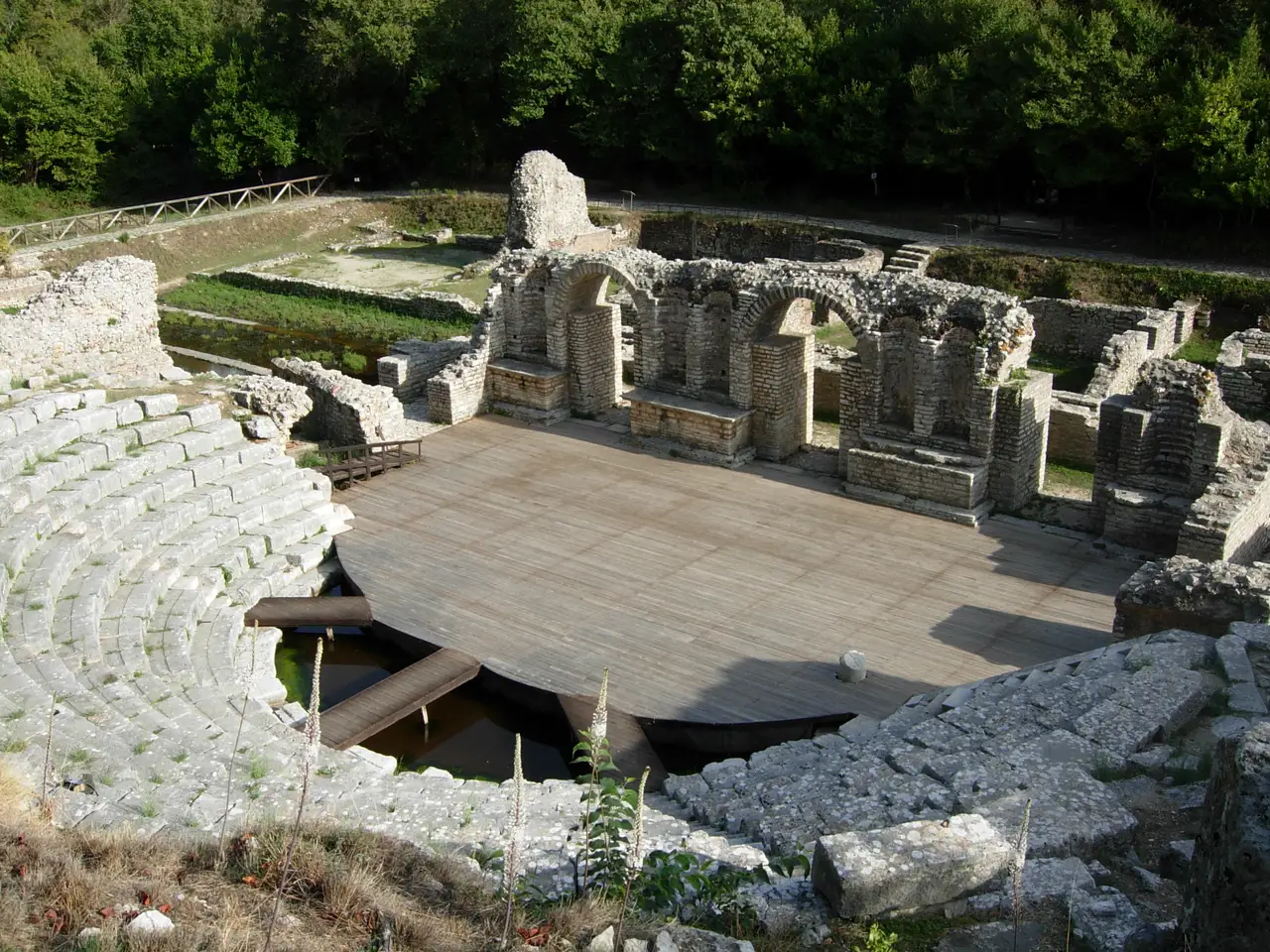 The ancient Greek theatre at Butrint with curved stone seating around a wooden stage floor and ruined stage buildings