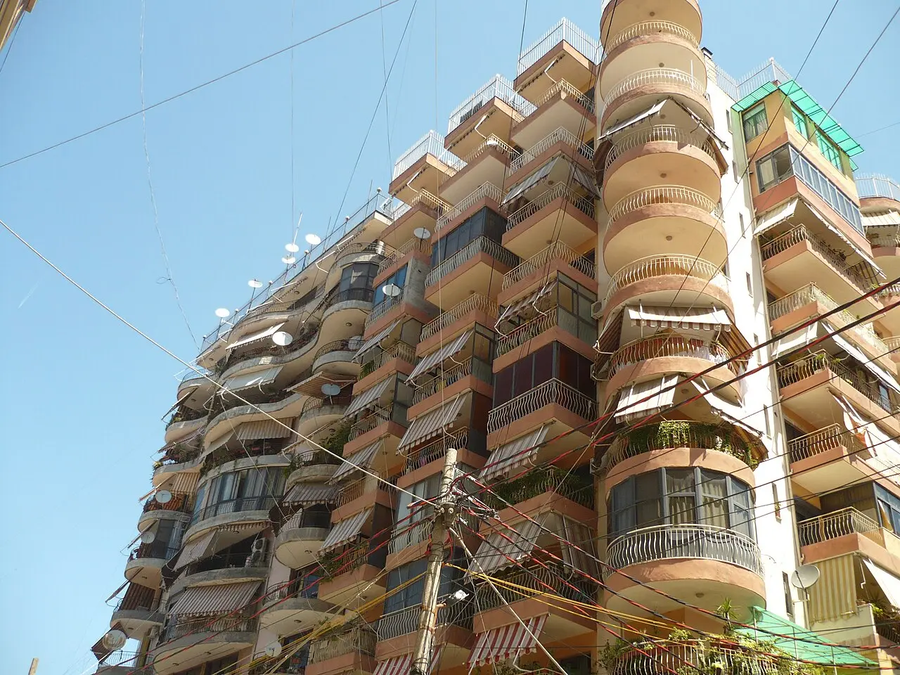 A tall Tirana apartment building with rounded balconies, awnings and overhead wires against a blue sky
