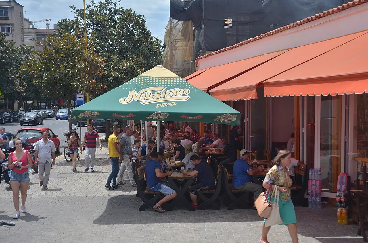 People sitting at outdoor cafe tables under a large umbrella on a sunny Tirana street