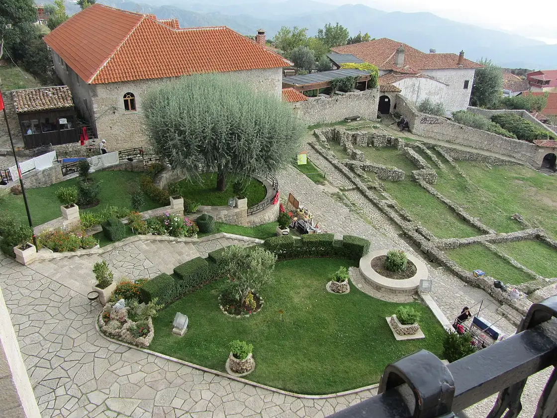 The green courtyard and old stone buildings inside Kruja Castle, with mountains behind