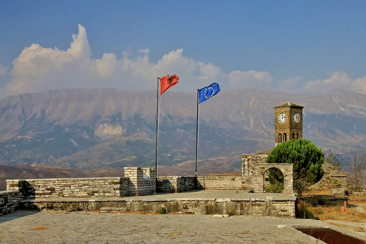The stone clock tower and the Albanian and EU flags on a terrace of Gjirokastër castle with mountains behind