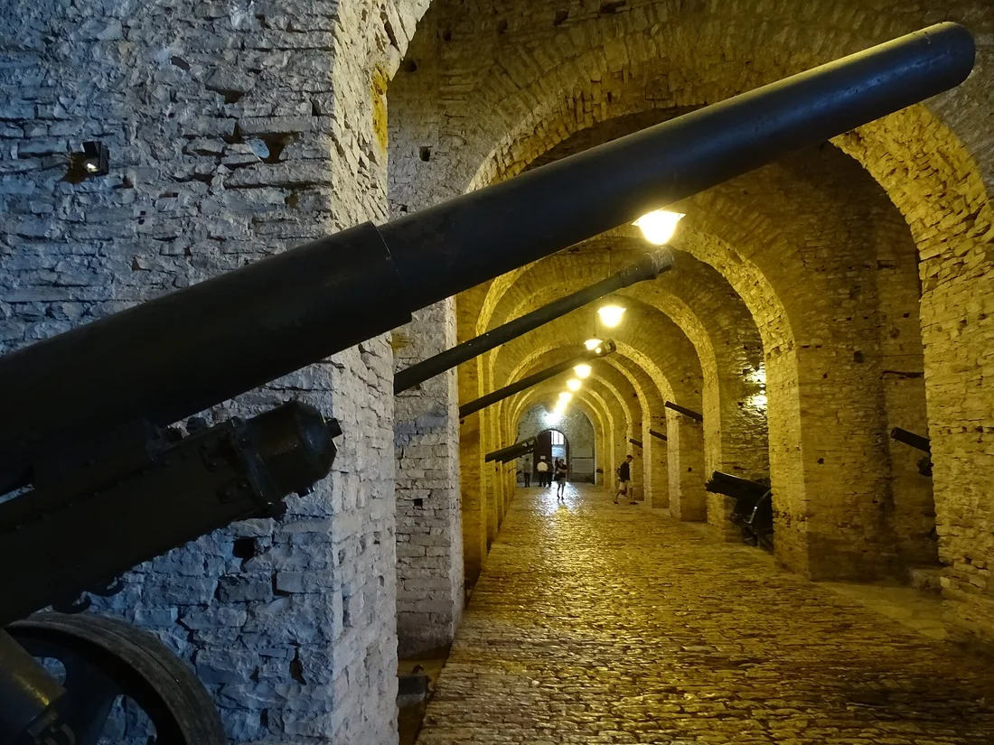 Vaulted stone gallery lit by lamps with old artillery barrels along the wall