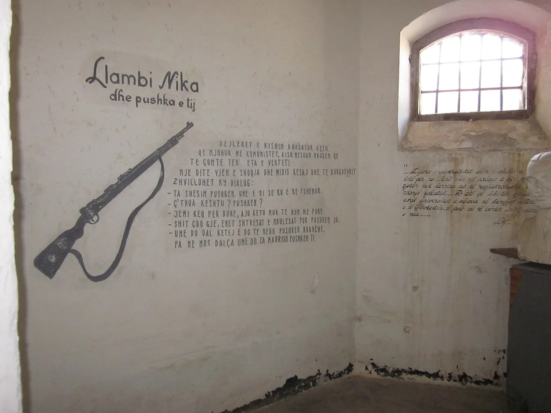 Whitewashed prison cell with a barred window and a rifle and text painted on the wall