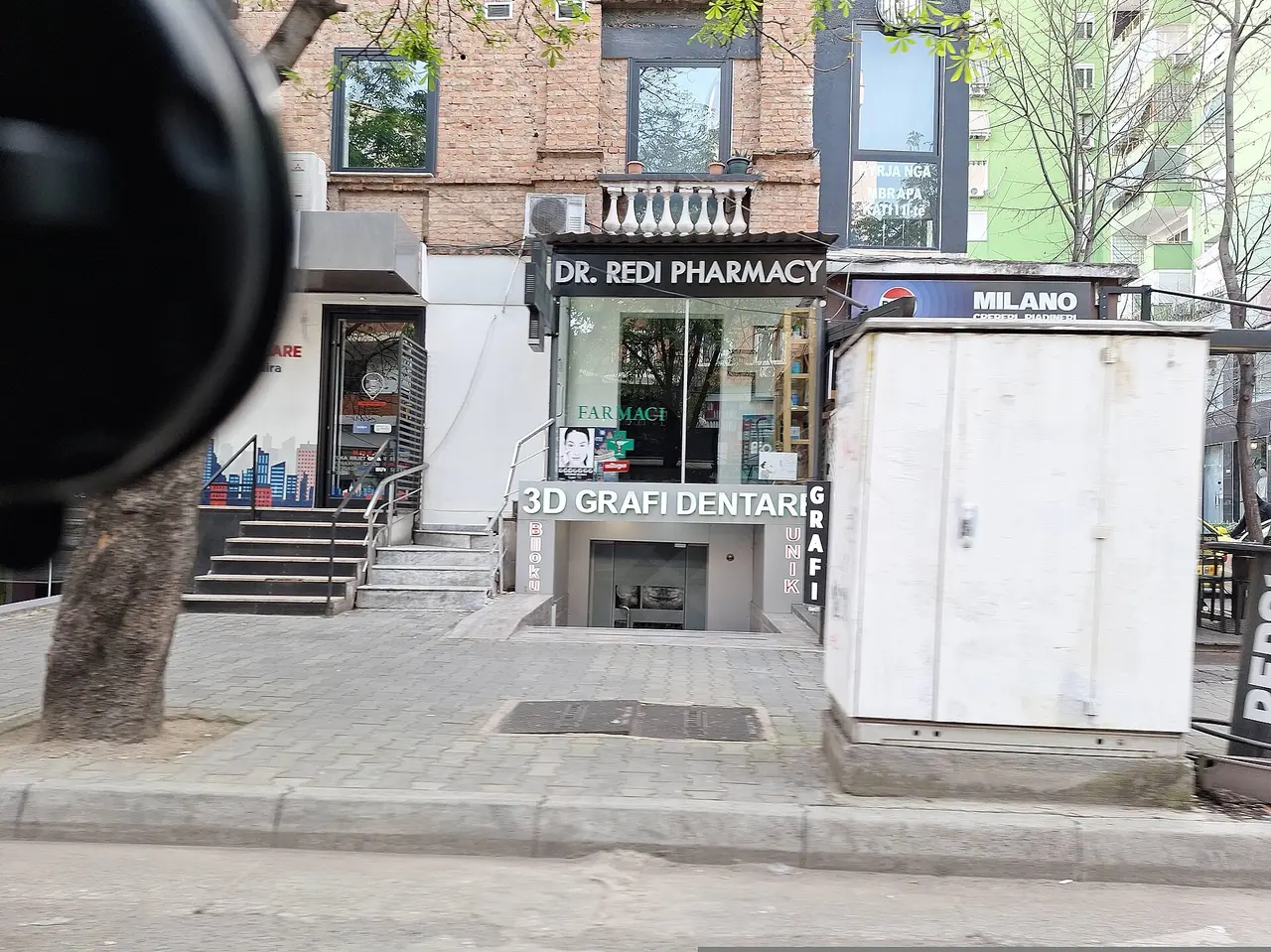 Street-level pharmacy frontage on a Tirana pavement with parked cars and apartment balconies