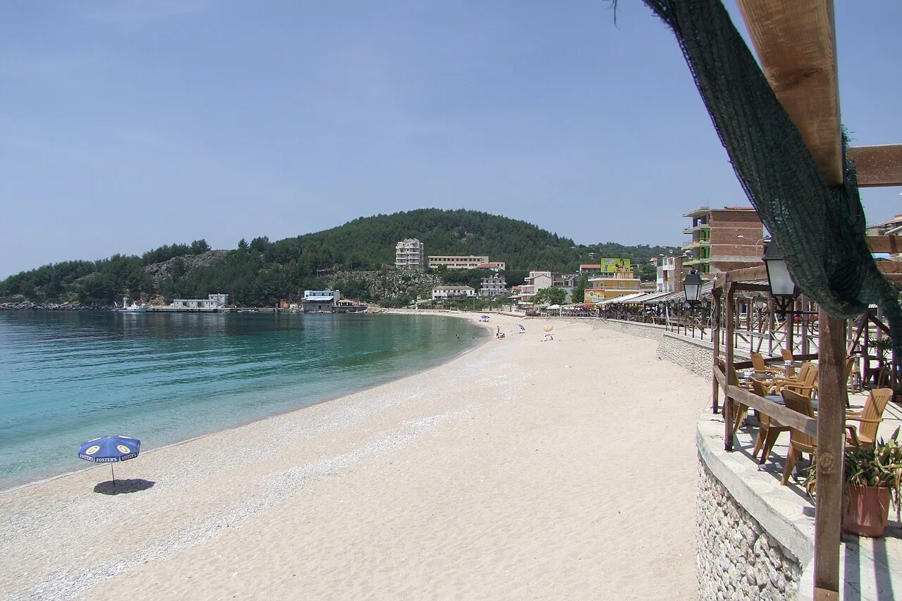 Himara's town beach of white pebbles curving to a headland, with waterfront buildings and a taverna terrace behind
