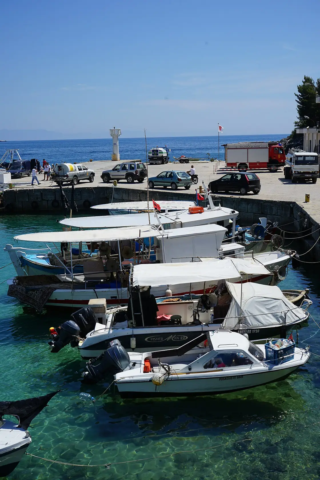 Fishing boats moored in the clear turquoise water of Himara harbour, with a small lighthouse on the quay