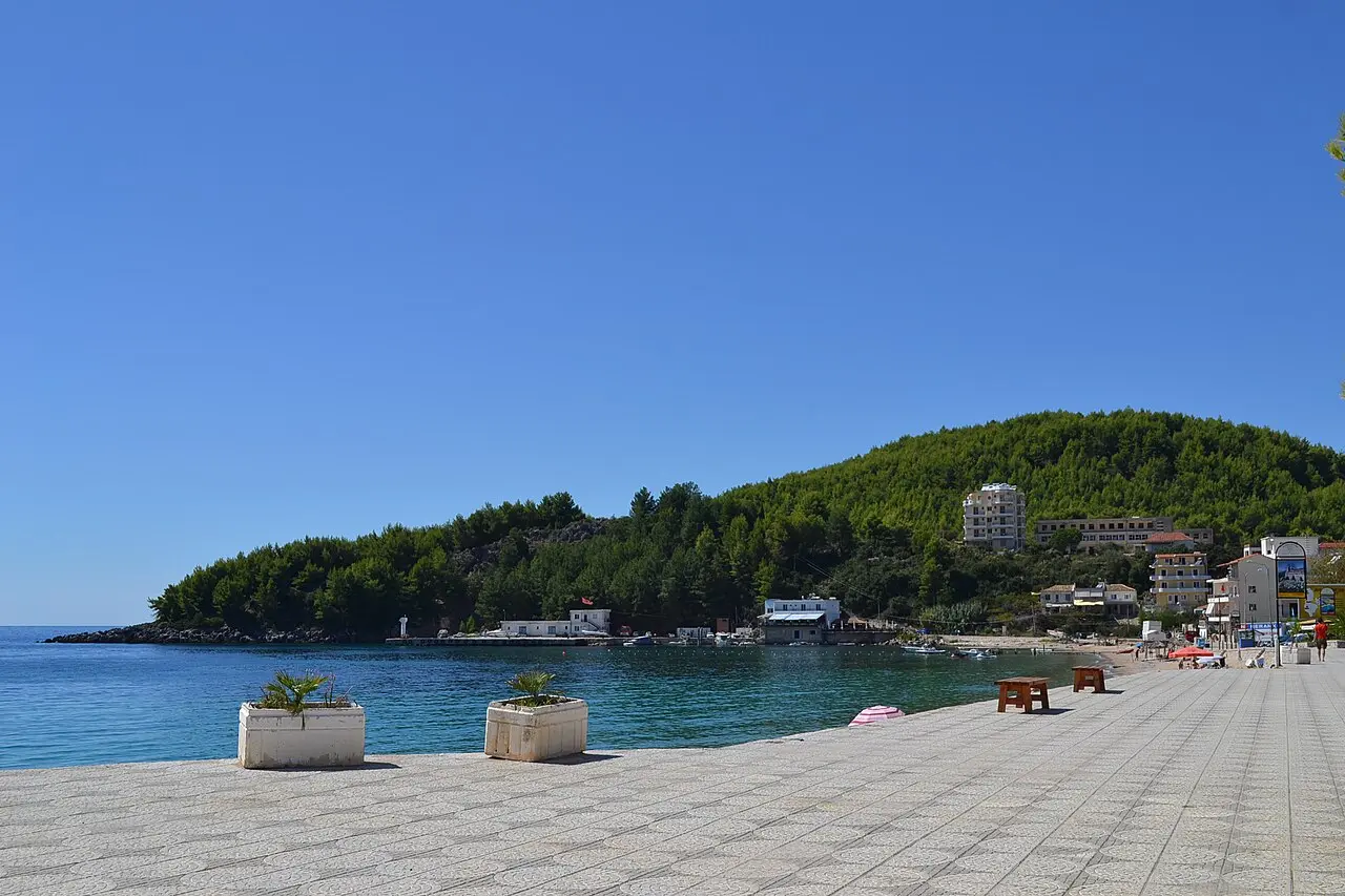 A quiet stone promenade beside a calm cove backed by a pine-covered headland near Himara