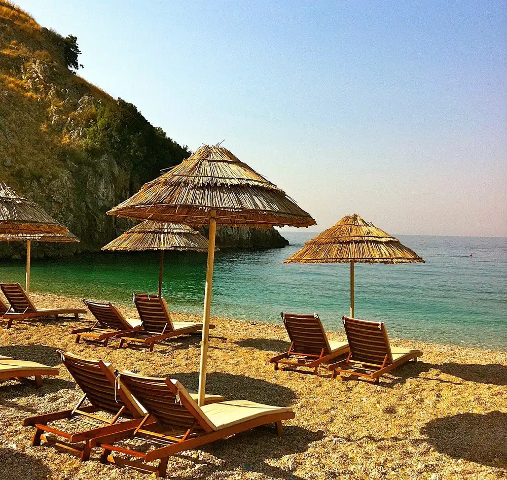 Thatched umbrellas and wooden sunbeds on a Himara pebble cove with clear turquoise water and a rocky headland