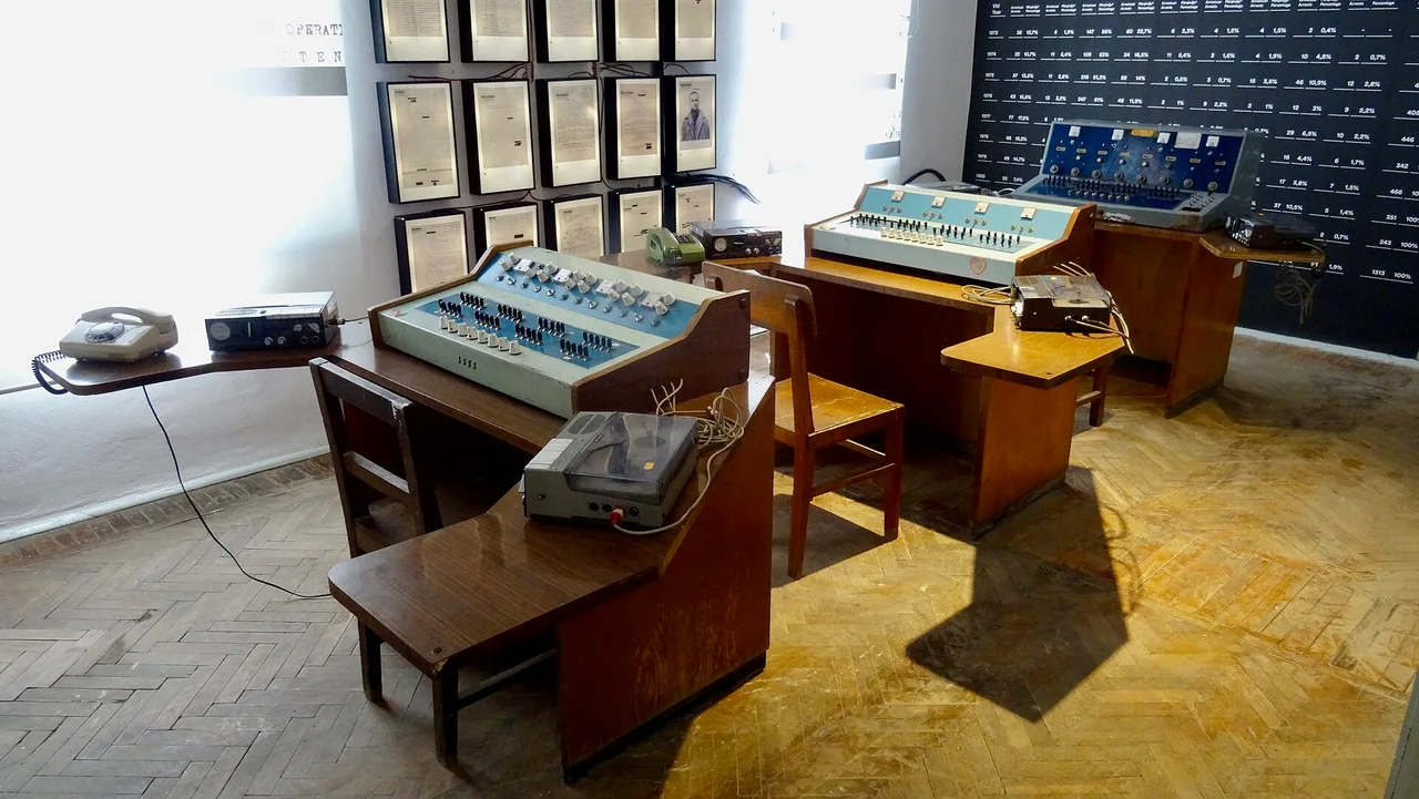 Blue listening consoles and reel tape recorders on wooden desks over a parquet floor, with framed documents on the wall