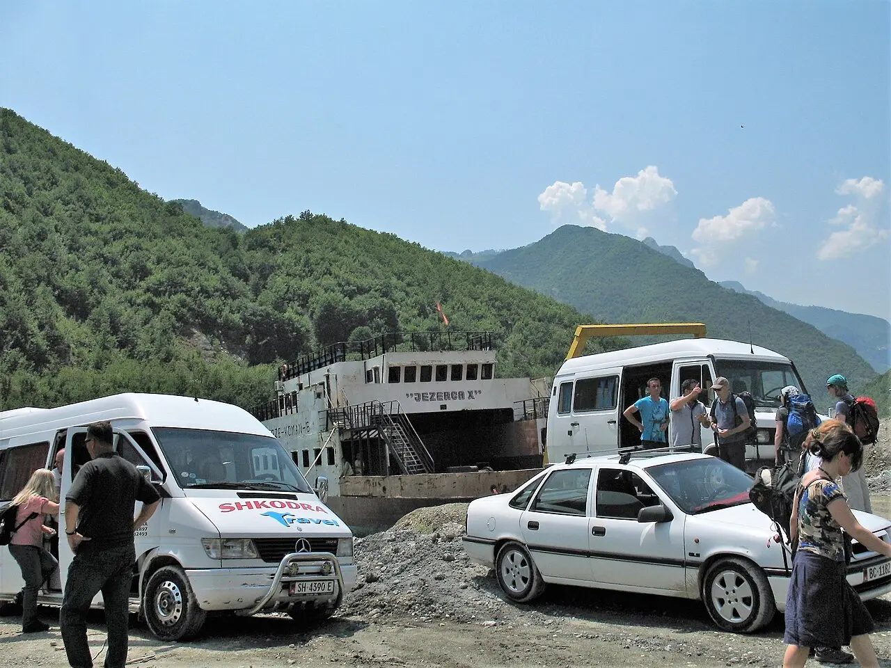 A car ferry loading minibuses and cars at the shore of Lake Koman, with green mountains rising behind