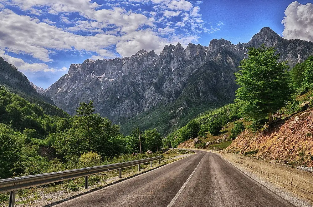 A mountain road curving through the Valbona Valley with jagged peaks of the Albanian Alps behind