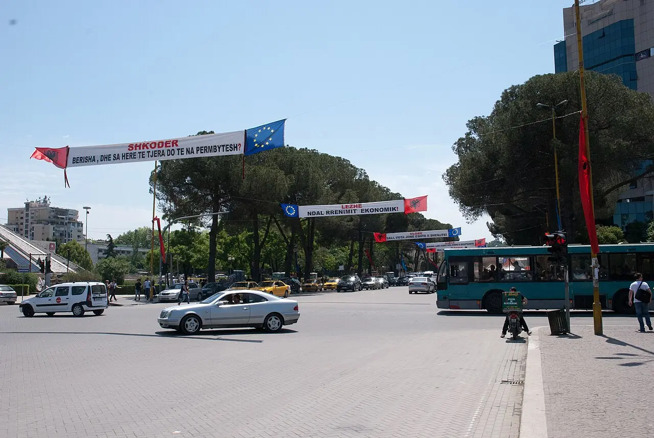 A busy junction in central Tirana with cars, a city bus and pedestrians crossing on a sunny day