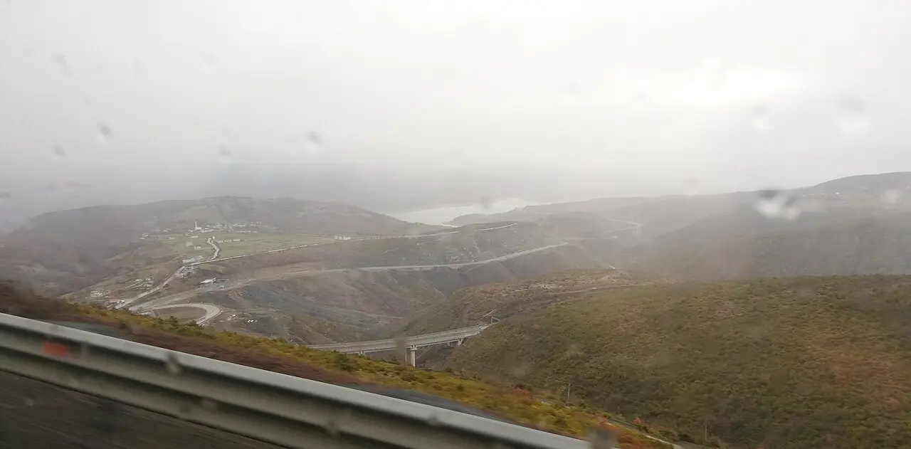 A winding two-lane road descending through misty Albanian hills, seen through a rain-spattered car window