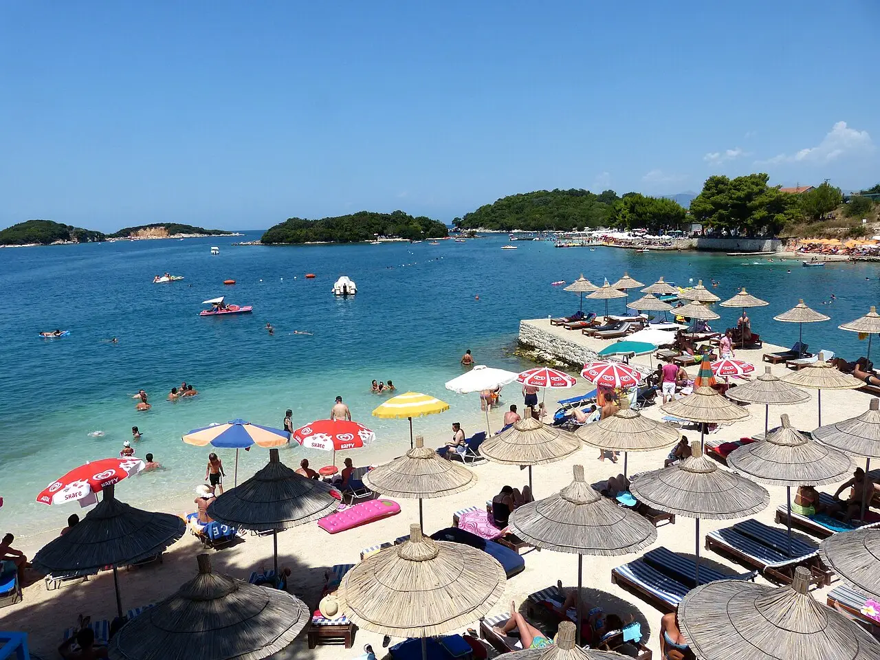 A busy Ksamil beach with straw sunshades, sun loungers and swimmers in the turquoise bay