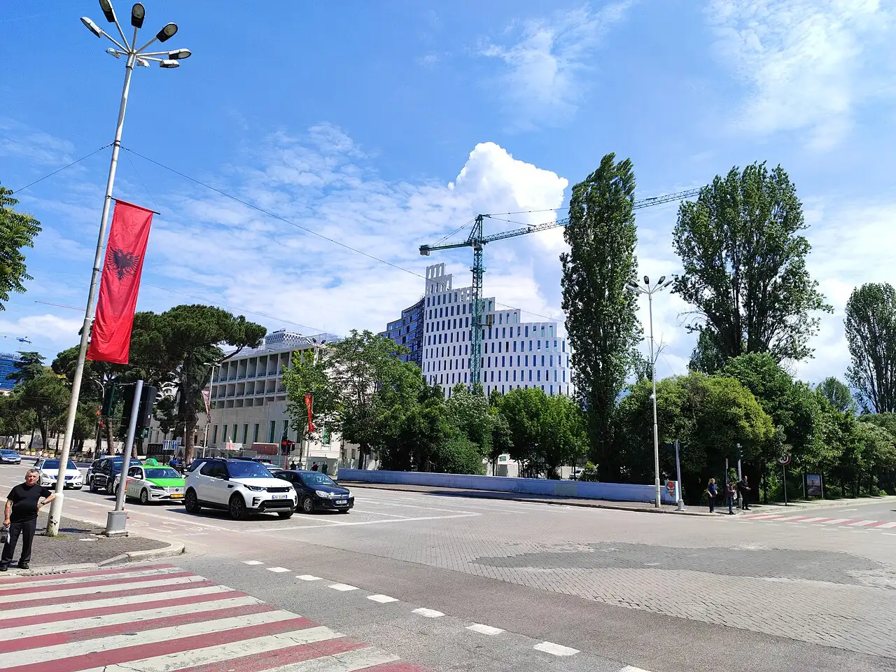 A quiet street crossing in central Tirana with an Albanian flag, a marked crosswalk and a modern office building behind trees