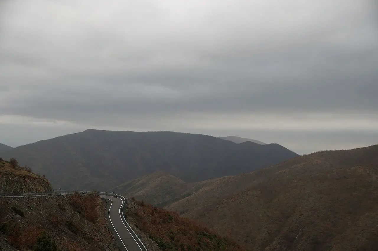 A winding mountain road with a crash barrier cutting through the hills of central Albania under a grey sky