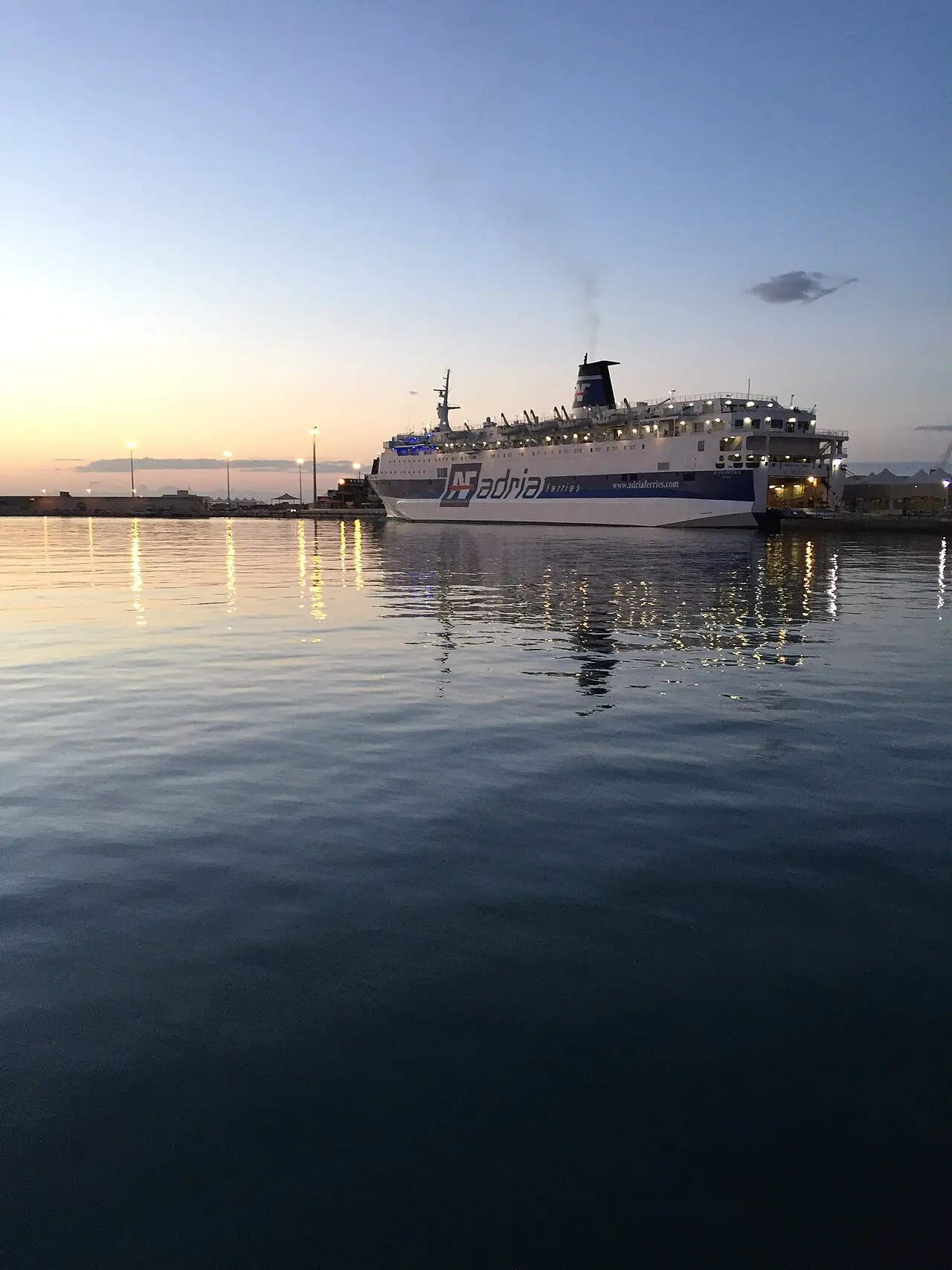 An Adria Ferries ship berthed at the port of Bari in Italy at dusk, lights reflected in calm water