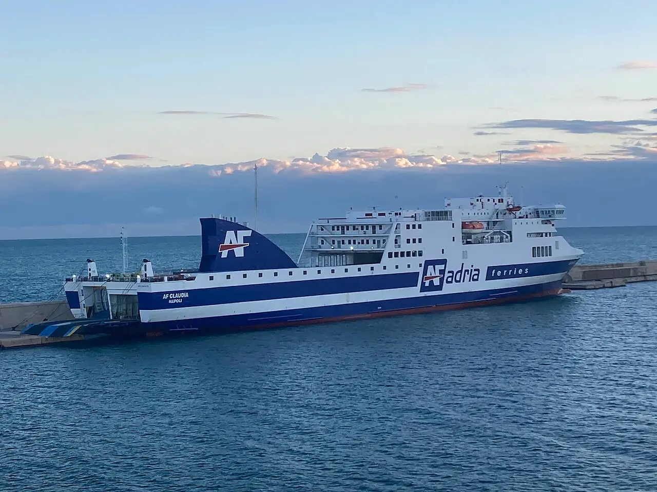 An Adria Ferries ship with its stern vehicle ramp lowered at a port quay, ready to load cars
