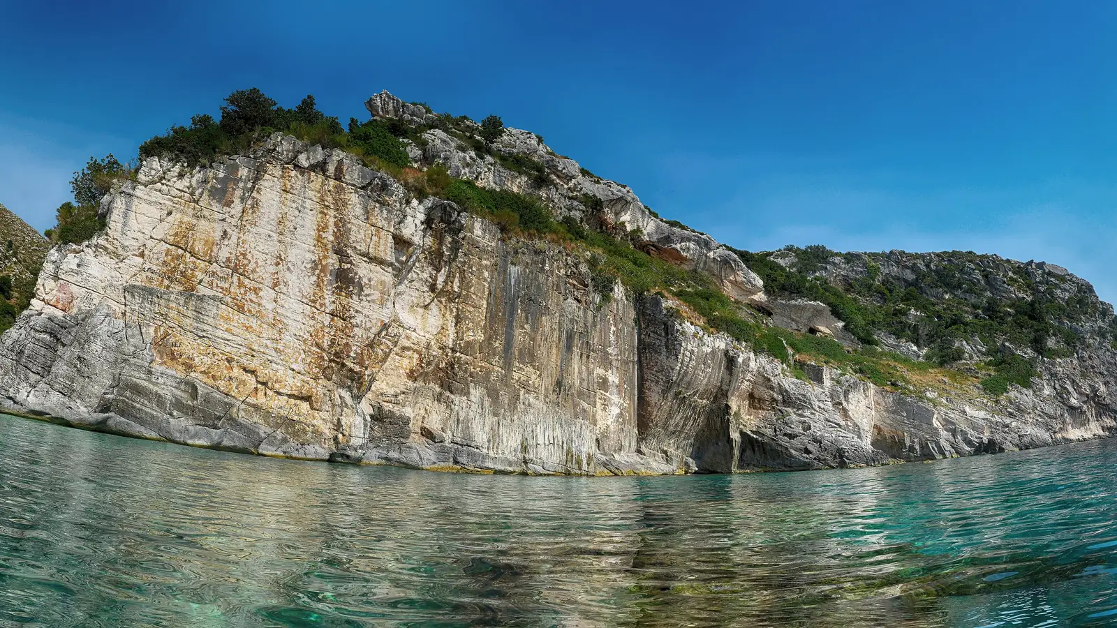 Pale limestone cliff at Grama Bay dropping into clear turquoise water, its face marked with panels of ancient carved inscriptions
