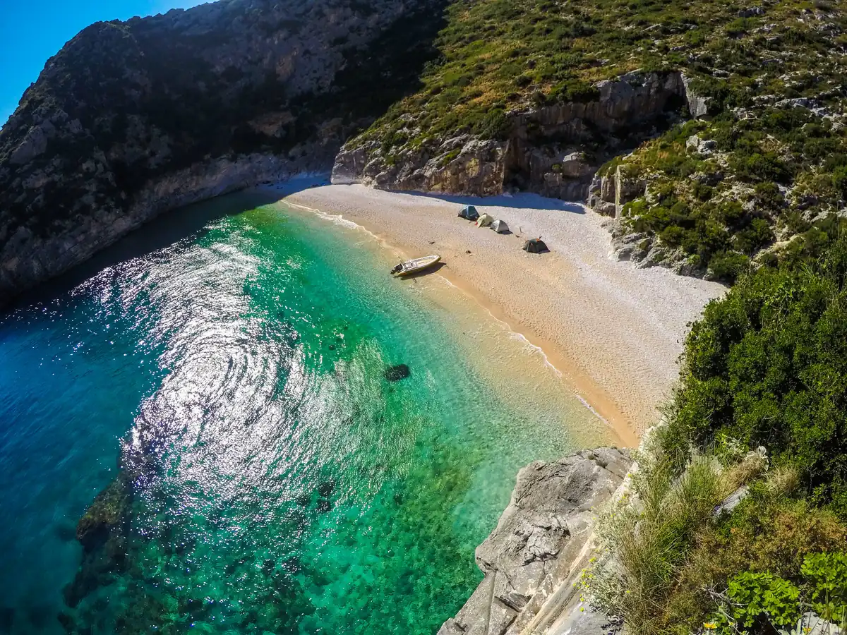 Aerial view of a white pebble beach and green water in a cove at Grama Bay with small tents on the shore