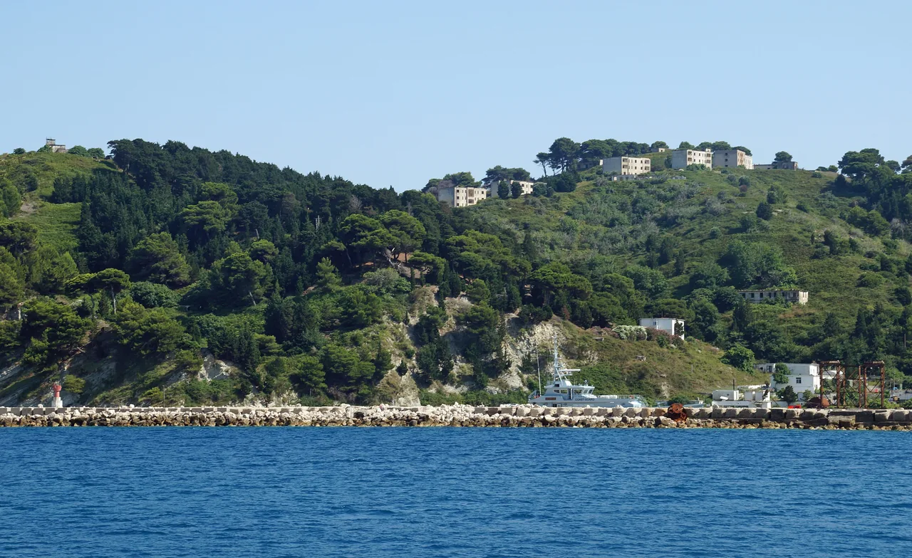 Abandoned apartment blocks above the small harbour on Sazan island with a patrol boat at the quay