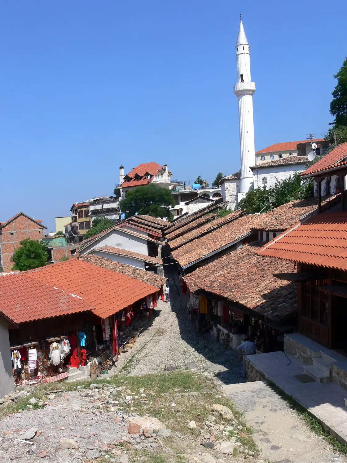 An elevated view down the two rows of red-tiled wooden bazaar shops in Kruja, with the cobbled lane between them and a white minaret rising above the old town